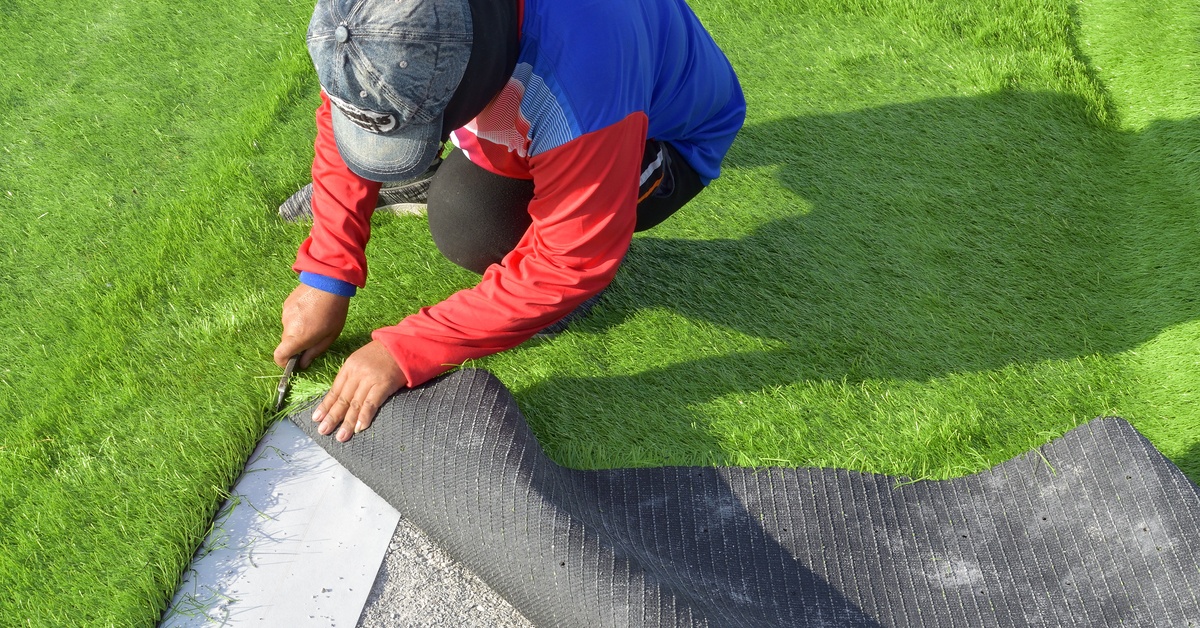 A person sitting on a green artificial turf field, using a cutting tool to separate it into large strips.