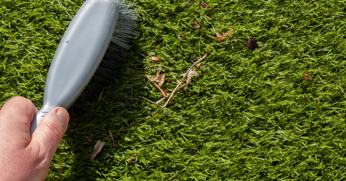 A hand holding a gray brush while sweeping small pieces of debris from bright green artificial turf.