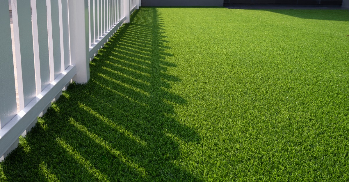 A white fence casting striped shadows across bright green artificial turf beside a home exterior and wall.