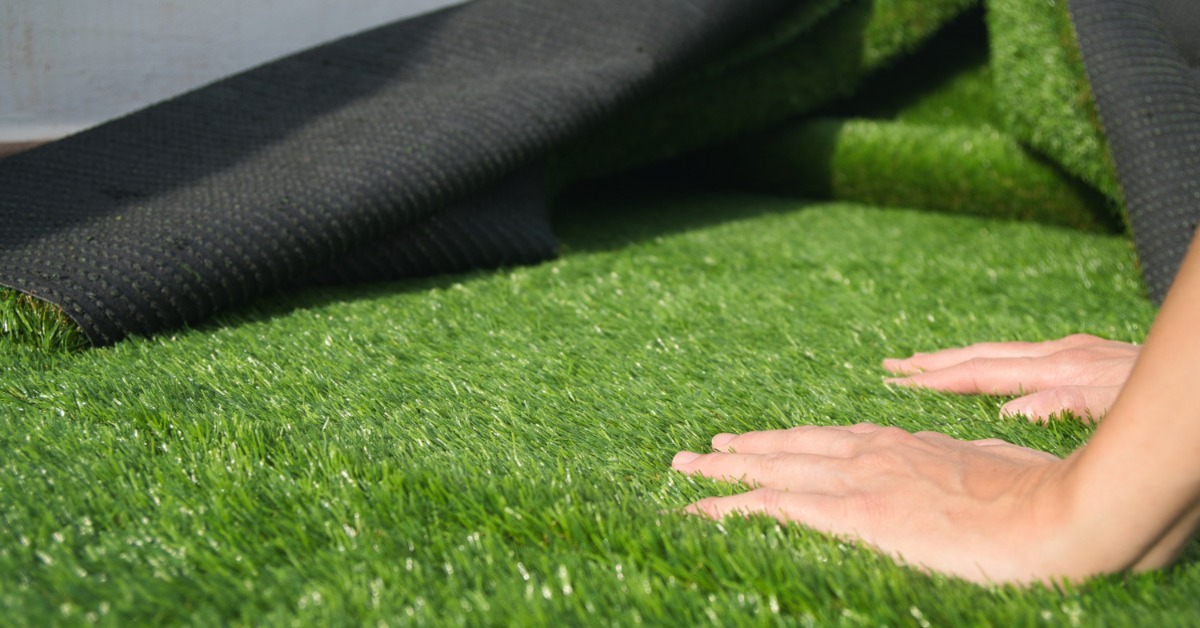 Hands pressing green artificial grass while a black backing layer is folded over the turf during installation.