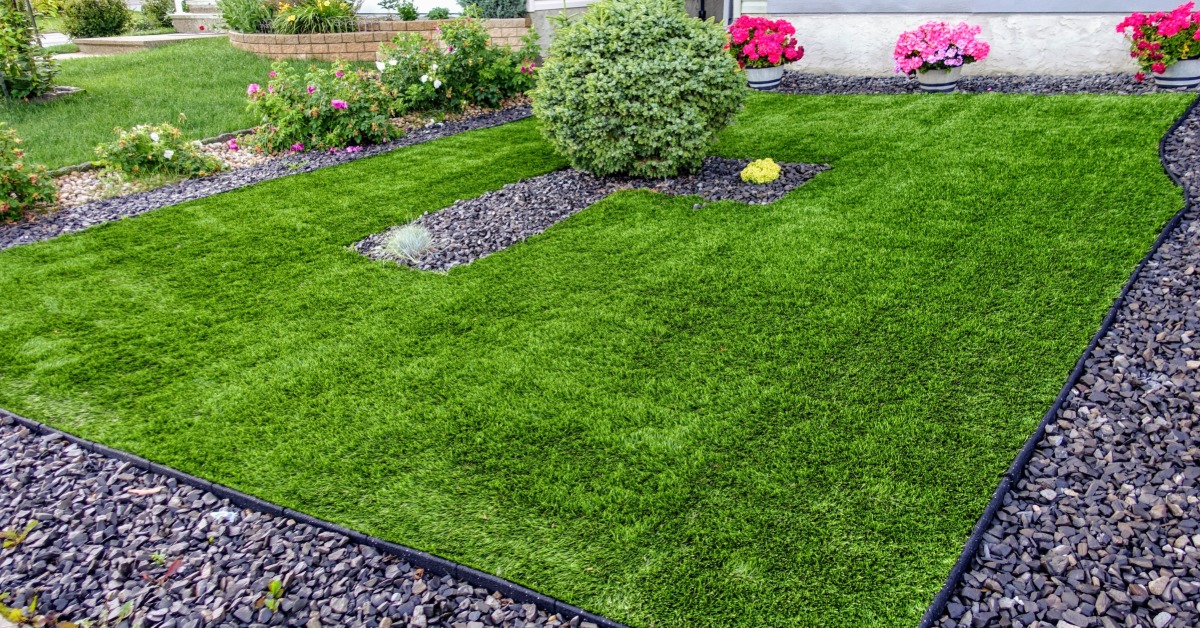 A green front yard lawn bordered by dark gravel, shrubs, pink flowers, and part of a house exterior.