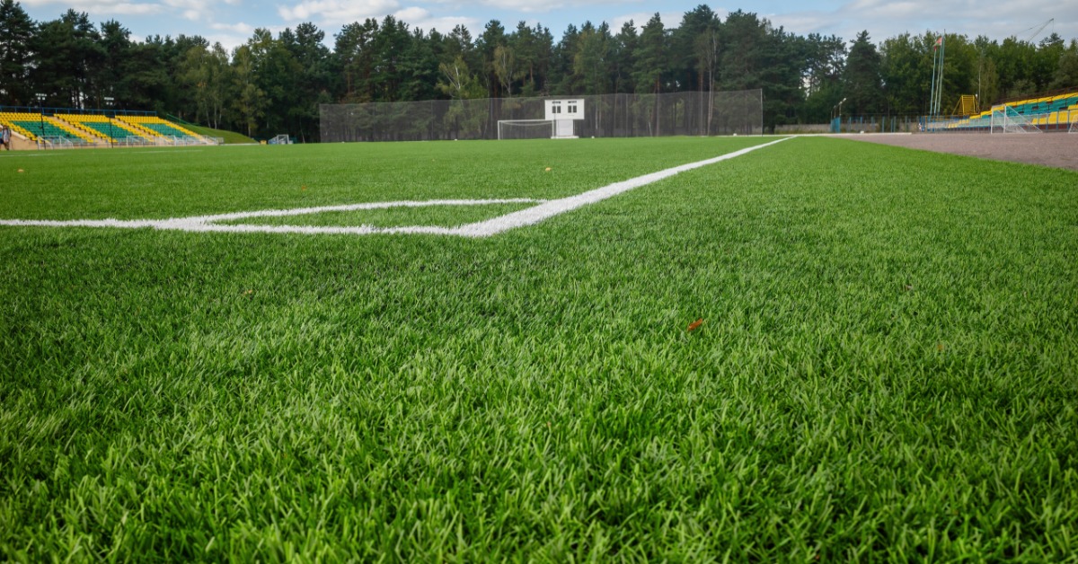 Green artificial turf with white field lines stretching across an empty stadium with trees in the background.