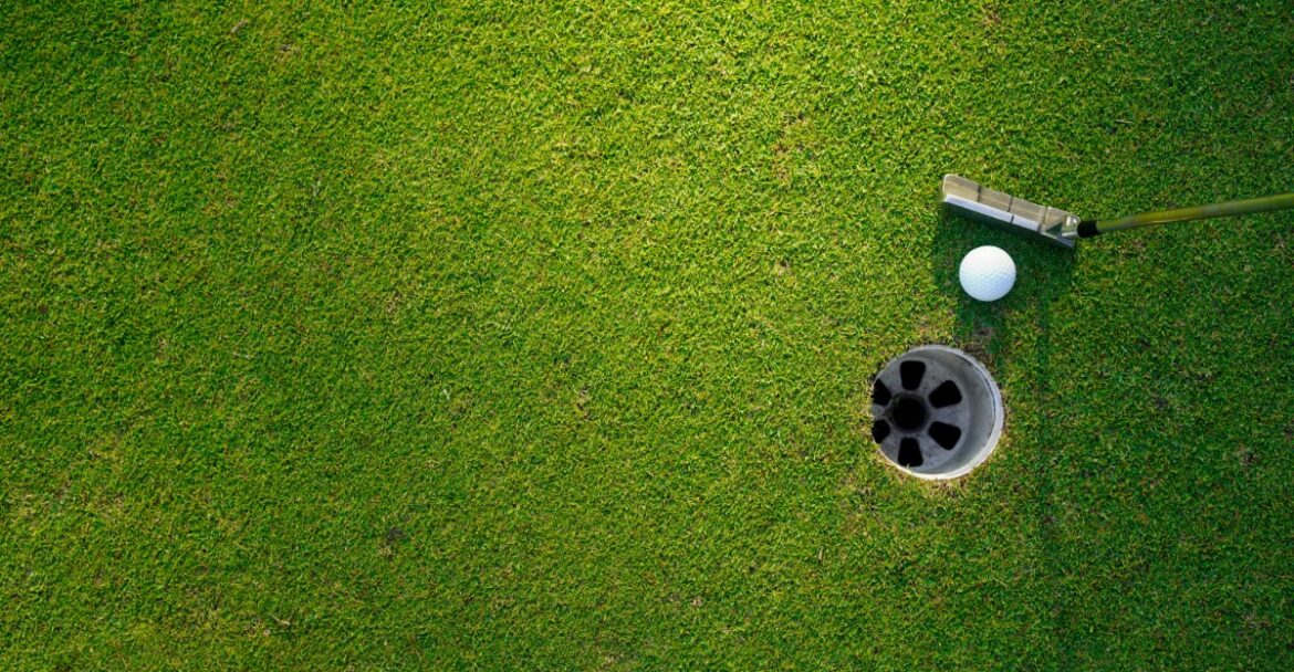 A white golf ball and putter resting near a circular hole on bright green turf with open grass around them.