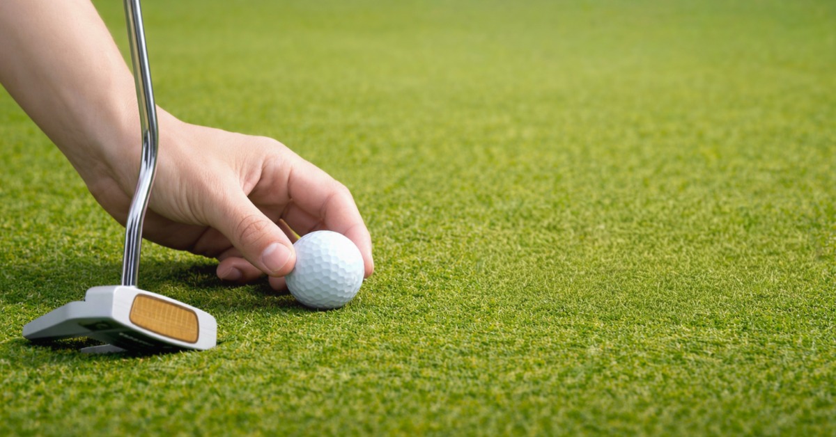A hand placing a white golf ball beside a putter on smooth green turf with open grass extending behind.