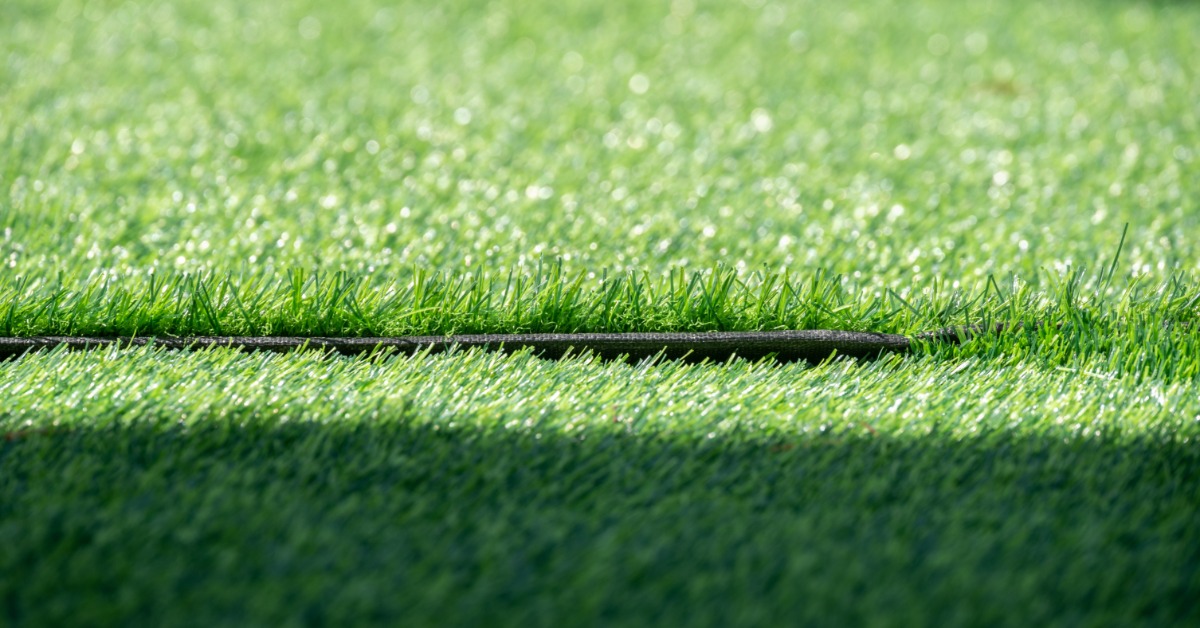 A green artificial turf surface with a narrow seam running horizontally between raised sections of grass.