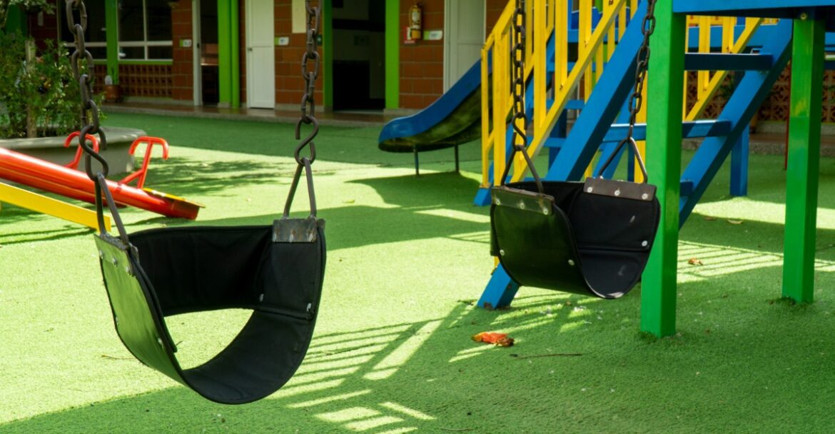 Two black rubber swings hanging over green turf near a colorful playground structure and slide in sunlight.