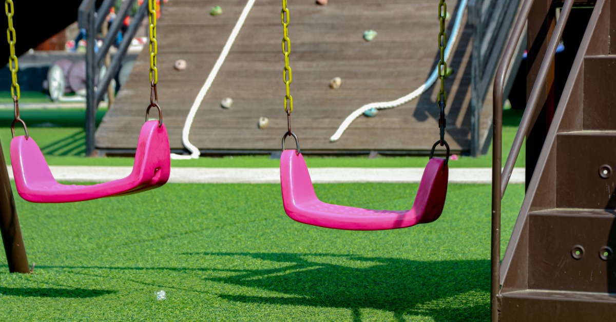 Two pink swings hanging over green turf beside a brown climbing structure with shadows across the grass.