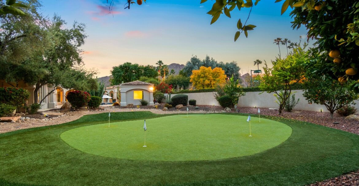 A backyard putting green with several flagsticks, surrounded by trees, shrubs, rocks, and neighboring buildings.