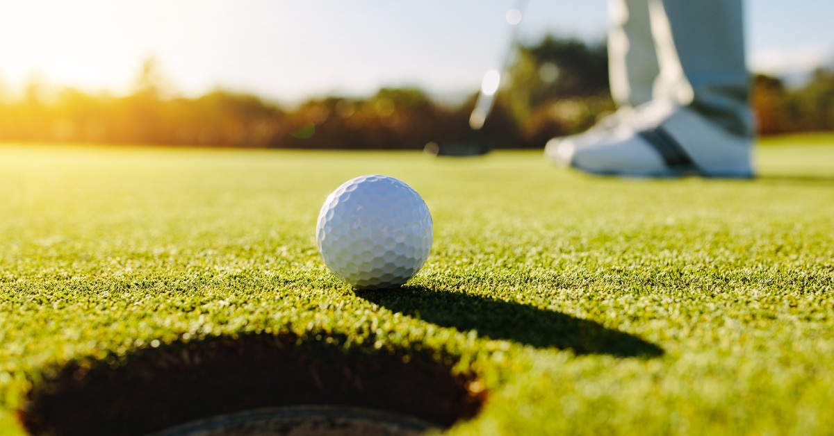A white golf ball resting near a hole on a putting green, with a person holding a club and standing in the background.
