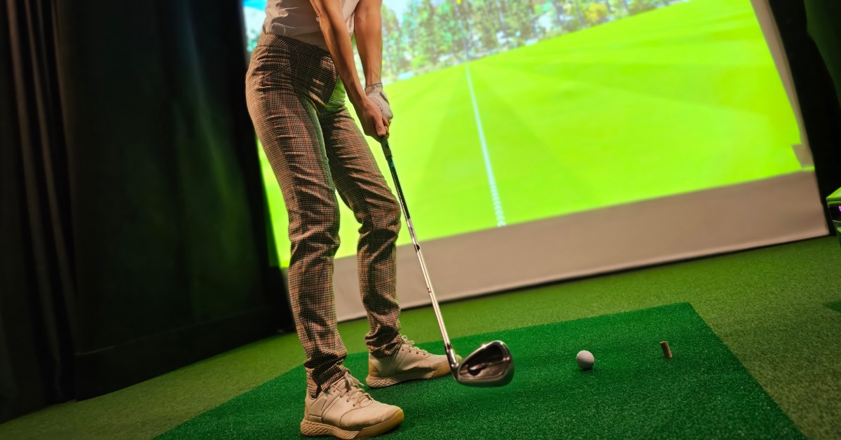 A person holding a golf club beside a ball on indoor turf, with a large golf course projection displayed behind them.